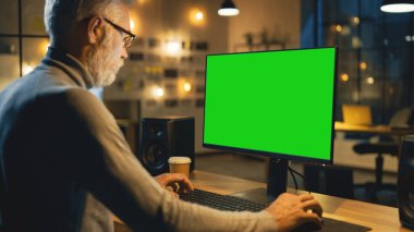 Masasında oturan yaratıcı Ortaçağ Tasarımcısı Yeşil Model Ekranlı Masaüstü Bilgisayarını Kullanıyor. Ofiste Şehir Penceresi Görünümü ile Bilgisayarlar üzerinde çalışan Akşam Yaratıcı Çalışanında