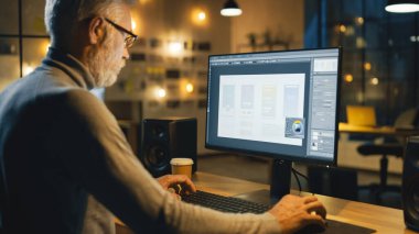 Masasında oturan yaratıcı Ortaçağ Mobil Yazılım Tasarımcısı Akıllı Telefon Uygulama Tasarım Süreci ile Masaüstü Bilgisayarını kullanır. Modern Stüdyo Ofisi