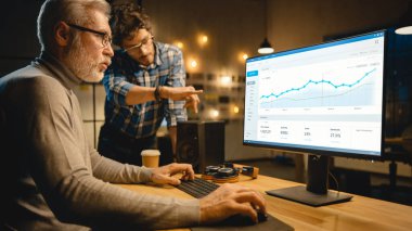 Profesyonel Üst Düzey İş Analisti Masaüstü Bilgisayarında Çizelgeler, Grafikler ve Ona Tavsiye Veren Genç Uzmanla Sohbet Ediyor. Akşamları Modern İş Ofisi