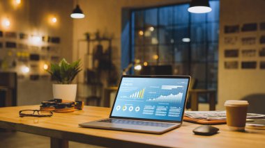 Ofisteki Masadaki Dizüstü bilgisayar Şirket Gelişim Verilerini Grafikler, Grafikler ve Yazılım UI ile gösteriyor. Arkaplanda Modern Ofis Stüdyosunda Büyük Şehir Penceresi ile Akşam