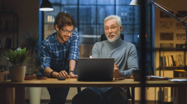 Yakışıklı Orta Çağ Girişimcisi ve Başarılı İşadamı Masasında oturan Laptop 'ta çalışıyor, Genç Meslektaşlarıyla Tartışıyor. Gülümsüyorlar ve eğleniyorlar. Akşamın geç saatlerinde şık bir ofis.
