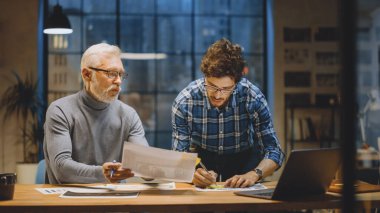 Baş Yaratıcı Tasarımcı Genç Geliştiriciyle Tasarım Çarşaflarını Tartışıyor. Tartışan iki profesyonel, belgelerle çalışan, doğru kavramı seçen. Modern Stüdyo Ofisi 'nde akşam