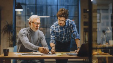 Baş Yaratıcı Tasarımcı Genç Geliştiriciyle Tasarım Çarşaflarını Tartışıyor. Tartışan iki profesyonel, belgelerle çalışan, doğru kavramı seçen. Modern Stüdyo Ofisi 'nde akşam