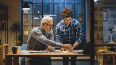 Baş Yaratıcı Tasarımcı Genç Geliştiriciyle Tasarım Çarşaflarını Tartışıyor. Tartışan iki profesyonel, belgelerle çalışan, doğru kavramı seçen. Modern Stüdyo Ofisi 'nde akşam