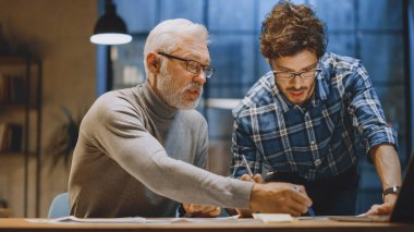 Baş Yaratıcı Tasarımcı Genç Geliştiriciyle Tasarım Çarşaflarını Tartışıyor. Tartışan iki profesyonel, belgelerle çalışan, doğru kavramı seçen. Modern Stüdyo Ofisi 'nde akşam