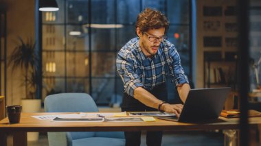 Masasında Çalışan Genç Yaratıcı Tasarımcı, Dizüstü bilgisayar ve Dockuments, Doğru Tasarım, Konsept ve Çizimler Üzerine Odaklandı. Şehir manzarası Pencere Görünümlü Arkaplan Akşam Ofisi Stüdyosunda
