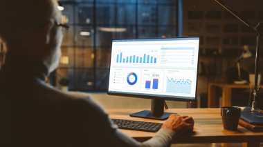 Masasında İstatistik, Çeşitli Veriler, Grafikler ve Grafiklerle Masaüstü Bilgisayarını kullanarak oturan kendinden emin orta yaşlı iş adamı üzerinde. Şehir Penceresi Görünümlü Şık Ofis Stüdyosunda Akşam