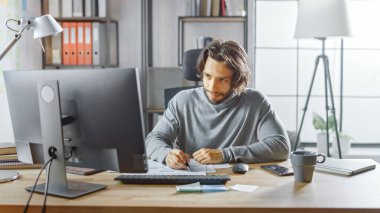 Yakışıklı Uzun Saçlı Girişimci Ofis 'teki Masasında Masaüstü Bilgisayarında Çalışıyor, Belgelerle Çalışıyor, Çizelgeleri Düzeltiyor, Grafikler Çiziyor, İstatistik ve Güncelleme Şirketi Stratejisi