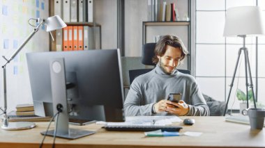 Yakışıklı Uzun Saçlı Girişimci Ofis 'teki Masasında Masaüstü Bilgisayarında, Belgeler ve Grafiklerle Çalışıyor. Akıllı Telefon, Sosyal Medya Uygulaması, E- posta Yazma, Mesajlaşma