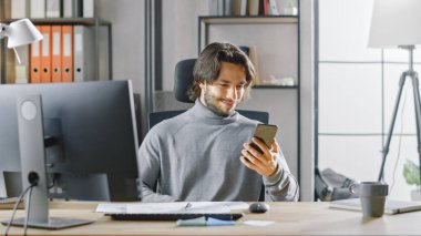 Yakışıklı Uzun Saçlı Girişimci Ofis 'teki Masasında Masaüstü Bilgisayarında, Belgeler ve Grafiklerle Çalışıyor. Akıllı Telefon, Sosyal Medya Uygulaması, E- posta Yazma, Mesajlaşma