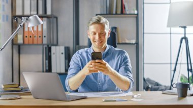 Çalışma masasında oturan yaratıcı girişimci, şık ofisteki bir dizüstü bilgisayarda çalışıyor, Sosyal Medya Uygulamaları için akıllı telefon kullanıyor, e-posta şirketleri, Haberler okuyor, İnternet 'e göz atıyor