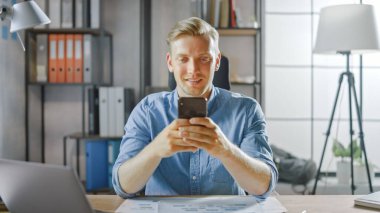 Çalışma masasında oturan yaratıcı girişimci, şık ofisteki bir dizüstü bilgisayarda çalışıyor, Sosyal Medya Uygulamaları için akıllı telefon kullanıyor, e-posta şirketleri, Haberler okuyor, İnternet 'e göz atıyor