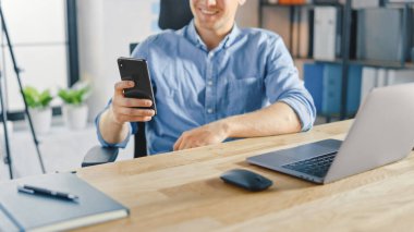 Çalışma masasında oturan yaratıcı girişimci, şık ofisteki bir dizüstü bilgisayarda çalışıyor, Sosyal Medya Uygulamaları için akıllı telefon kullanıyor, e-posta şirketleri, Haberler okuyor, İnternet 'e göz atıyor