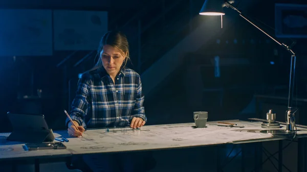 La ingeniera sentada en su escritorio funciona con planos colocados en una mesa, utiliza lápiz, regla y tableta digital. En las instalaciones de ingeniería de diseño industrial oscuro —  Fotos de Stock