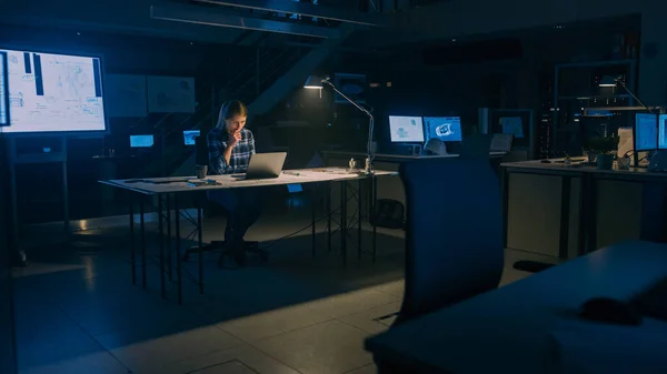 Linda engenheira sentada em sua mesa funciona em um computador portátil. Plantas deitadas numa mesa. Na Dark Industrial Design Engineering Facility. Lâmpada de luz quente — Fotografia de Stock