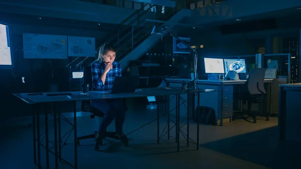 La hermosa ingeniera sentada en su escritorio trabaja en una computadora portátil. Planos acostados en una mesa. En las instalaciones de ingeniería de diseño industrial oscuro. —  Fotos de Stock