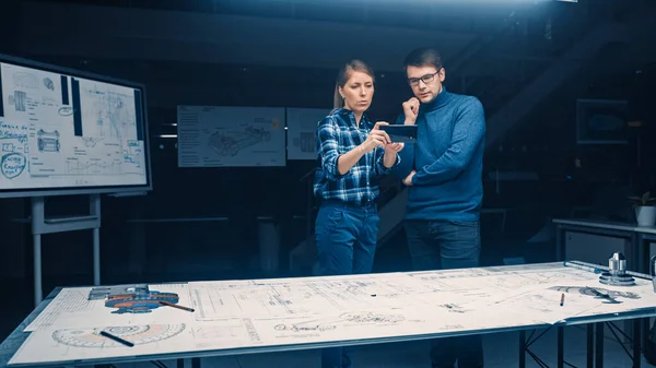 Dos jóvenes ingenieros de desarrollo de software hablan y usan teléfonos inteligentes con realidad aumentada para leer e interactuar con los planos del concepto de automóvil y motor que se encuentran en el escritorio — Foto de Stock