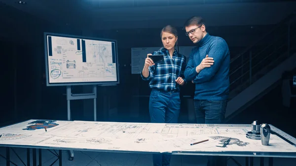 Два молодых инженера по разработке программного обеспечения говорят и используют смартфон с дополненной реальностью для чтения и взаимодействия с концепцией автомобиля и двигателя чертежи лежат на столе — стоковое фото