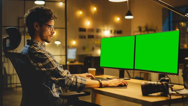 Tarde tarde en la oficina creativa: Fotógrafo profesional trabaja en una computadora de escritorio con dos pantallas simuladas verdes. Estudio moderno con bombillas colgantes — Foto de Stock