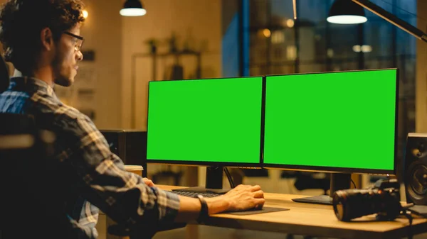 Over de schouder: creatieve ontwerper zit aan zijn bureau maakt gebruik van Desktop Computer met twee groene Mock-up schermen. Beroepsbureau Medewerker 's avonds laat aan het werk in zijn studio — Stockfoto