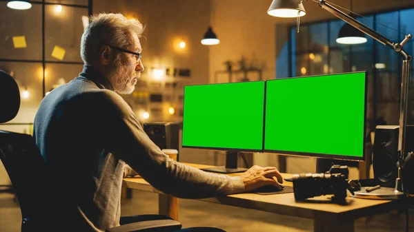 Kreatív középkorú tervező ül az asztalánál használ asztali számítógép két zöld Mock-up képernyők. Szakmai Office alkalmazott Késő estig dolgozik műtermében — Stock Fotó