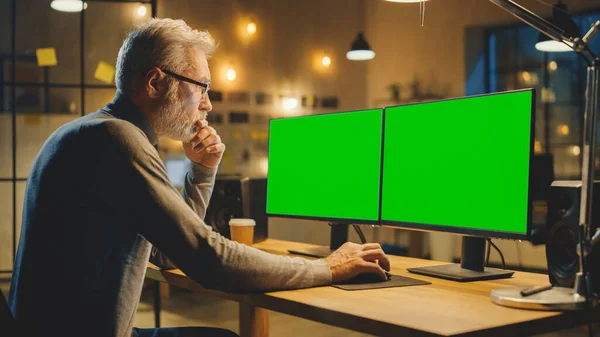 Δημιουργικός μεσήλικας σχεδιαστής που κάθεται στο γραφείο του χρησιμοποιεί επιτραπέζιο υπολογιστή με δύο πράσινες mock-up οθόνες. Επαγγελματίας υπάλληλος γραφείου που εργάζεται αργά το βράδυ στο στούντιο του — Φωτογραφία Αρχείου
