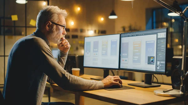 Diseñador creativo de software móvil de mediana edad sentado en su escritorio utiliza una computadora de escritorio con dos pantallas que muestran el proceso de diseño de aplicaciones para teléfonos inteligentes. Oficina de estudio moderno con estilo — Foto de Stock