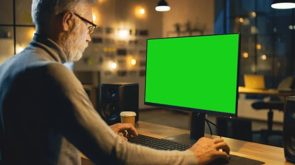 Diseñador creativo de mediana edad sentado en su escritorio utiliza una computadora de escritorio con pantalla verde simulada. Por la noche, Creative Employe trabaja en computadoras en la oficina con vista a la ventana de Cityscape —  Fotos de Stock