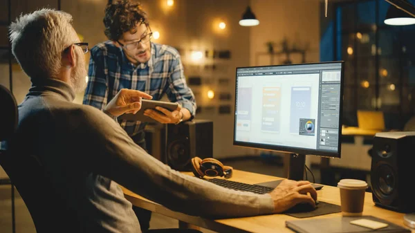 En la noche creativa Edad Media Diseñador de software móvil utiliza la computadora de escritorio con el diseño de aplicaciones Smartphone, Tiene conversación con el especialista más joven que utiliza Tablet Computer. —  Fotos de Stock