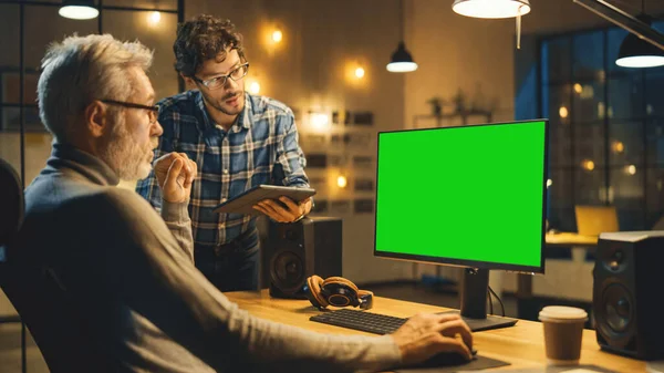 В вечернем творческом средневековье человек работает на настольном компьютерном зеленом макете экрана, беседует с более молодым специалистом, который использует планшетный компьютер. Стильный офис большого города в конце вечера — стоковое фото