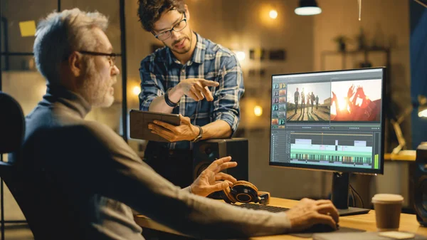 Evening in Creative Office: Professional Middle Aged Videographer Works on Desktop Computer, Has Conversation with Younger Specialist who Uses Tablet Computer. Modern Video Developers Office — Stock Photo, Image