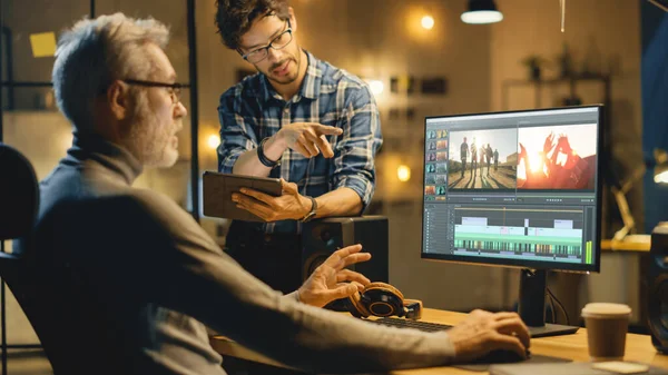 Soirée dans Creative Office : Vidéographe professionnel du Moyen Âge travaille sur ordinateur de bureau, a une conversation avec le jeune spécialiste qui utilise un ordinateur tablette. Bureau des développeurs vidéo modernes — Photo