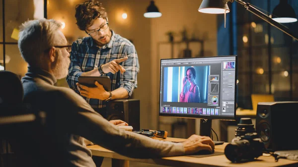 Dans la soirée créative Moyen Age Photo Editor fonctionne sur un ordinateur de bureau utilise un logiciel d'édition d'images, a une conversation avec le jeune spécialiste qui utilise Tablet Computer. Bureau élégant en soirée — Photo