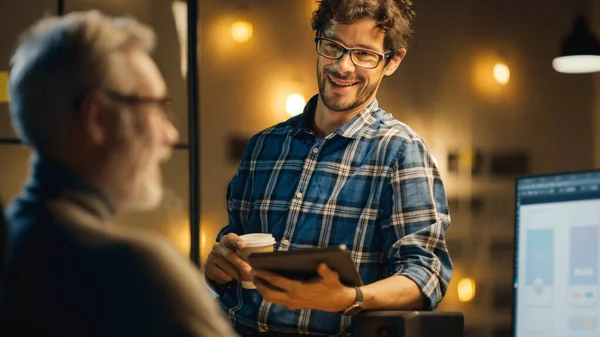 In the Evening Creative Middle Aged Smartphone Software Developer Works on a Desktop Computer with Screen show Application Design, Has Conversation with Younger Specialist who use Tablet Computer — 스톡 사진