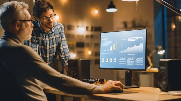 プロのシニアビジネスマーケットアナリストは、チャート、グラフを扱うデスクトップコンピュータ上で動作し、彼に助言を与える若い専門家と会話を持っています。夕方の近代的な営業所 — ストック写真