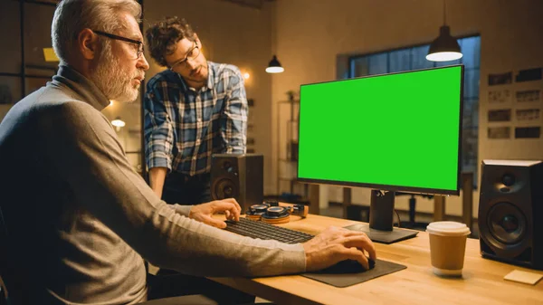 In de avond creatieve middelbare leeftijd Man werkt op een Desktop Computer Green Mock-up Screen, Heeft een gesprek met jongere specialist die Tablet Computer gebruikt. Stijlvol Big City Office laat in de avond — Stockfoto