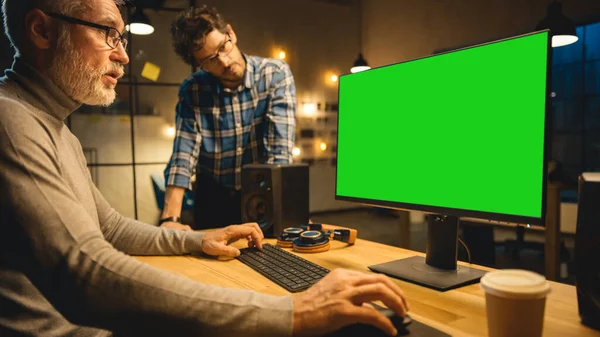 У Evening Creative Middle Aged Man працює на настільному комп'ютері Green Mock-up Screen, має розмову з молодшим спеціалістом, який використовує планшетний комп'ютер. Stylish Big City Office пізно ввечері — стокове фото