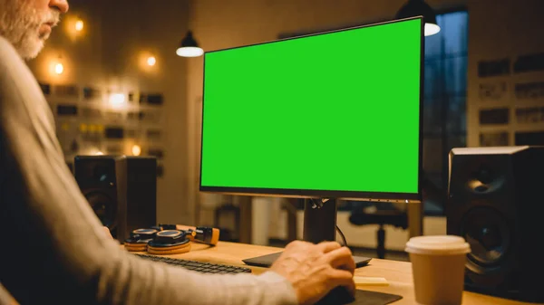 Diseñador creativo de mediana edad sentado en su escritorio utiliza la computadora de escritorio con dos pantallas simuladas verdes. Empleado profesional de oficina que trabaja hasta tarde en la noche en su estudio — Foto de Stock