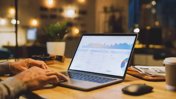 Nahaufnahme An den Händen des männlichen Spezialisten, der an einem Laptop arbeitet, zeigt der Bildschirm verschiedene Daten und Statistiken an. Im Hintergrund Abend im stilvollen Atelierbüro — Stockfoto