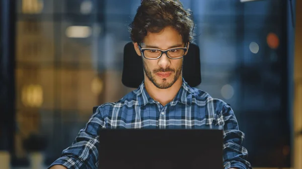 Portret przystojnego i udanego młodego przedsiębiorcy kreatywnego pracującego przy jego biurku przy użyciu laptopa. Praca z przytulnej pracowni Home Office z widokiem na miasto w nocy — Zdjęcie stockowe