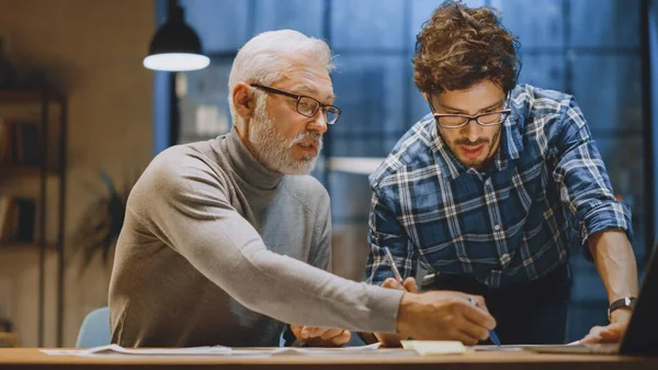 Baş Yaratıcı Tasarımcı Genç Geliştiriciyle Tasarım Çarşaflarını Tartışıyor. Tartışan iki profesyonel, belgelerle çalışan, doğru kavramı seçen. Modern Stüdyo Ofisi 'nde akşam