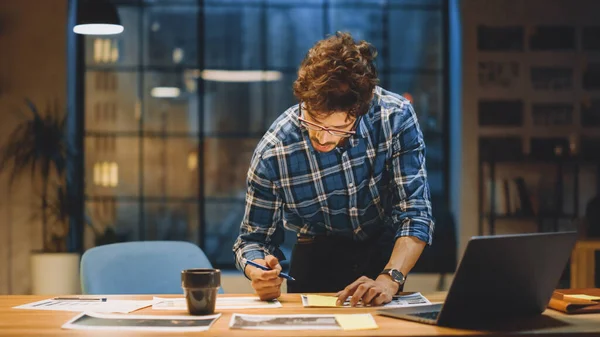 De jonge creatieve ontwerper werkt aan zijn bureau en concentreert zich op het papierwerk, het kiezen van het juiste ontwerp, concept en schetsen. In de achtergrond Avondkantoor Studio met Venster — Stockfoto