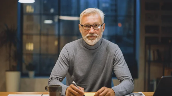 Retrato del apuesto y exitoso hombre de negocios barbudo de mediana edad que trabaja en su escritorio usando una computadora portátil. Sonrisas en la cámara. Trabajar de noche desde un acogedor estudio de oficina en casa con ventana — Foto de Stock