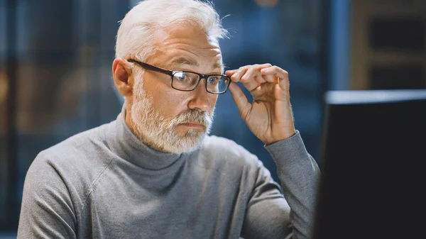 Retrato del apuesto y exitoso hombre de negocios barbudo de mediana edad que trabaja en su escritorio usando una computadora portátil y ajustando sus gafas. Trabajar de noche desde un acogedor estudio de oficina en casa con ventana — Foto de Stock