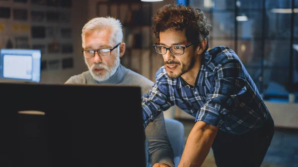 Diseñador creativo principal muestra la pantalla de la computadora de escritorio a un desarrollador joven, discuten el proyecto. Dos profesionales discutiendo. Elegir el concepto de diseño correcto. Noche en la oficina del estudio —  Fotos de Stock