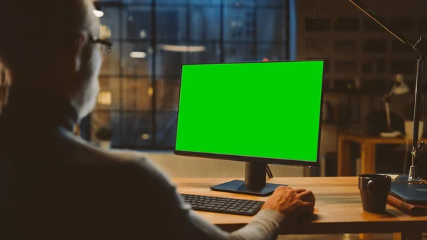 Over the Shoulder: Homem de meia-idade confiante sentado em sua mesa usando o computador desktop com tela verde falsa. Noite no elegante estúdio de escritório com vista para a janela da cidade — Fotografia de Stock