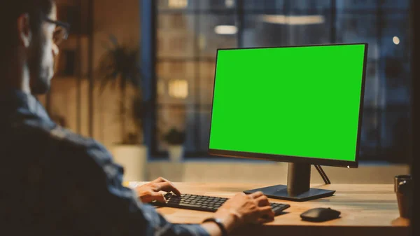 Over the Shoulder: Criativo jovem sentado em sua mesa usando o computador desktop com tela verde Mock-up. Noite no elegante estúdio de escritório com vista para a janela da cidade — Fotografia de Stock