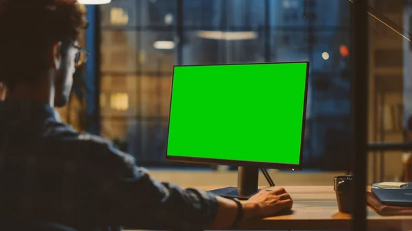 Über die Schulter: Kreativer junger Mann sitzt an seinem Schreibtisch und bedient sich eines Desktop-Computers mit grünem Bildschirm-Attrappe. Abend im stilvollen Büro-Studio mit Blick auf die Stadt — Stockfoto