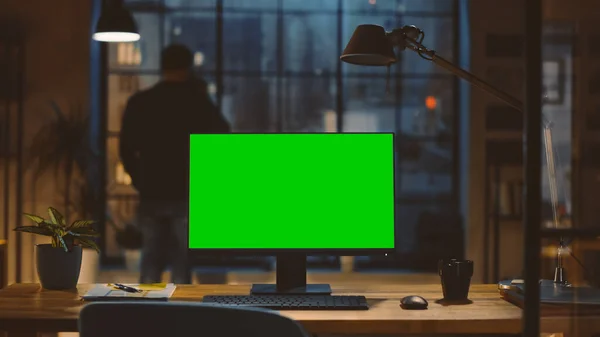Desktop-Computer mit grünem Bildschirm, der auf dem Schreibtisch im modernen Kreativbüro steht. Im Hintergrund blickt Designer Drinks aus einer Tasse aus dem Fenster auf die nächtliche Stadt. — Stockfoto