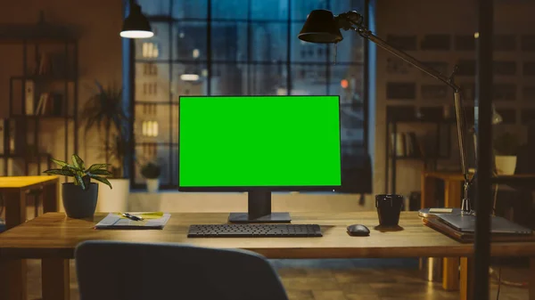 Computador de mesa com tela verde Mock-up em pé na mesa de madeira no escritório criativo moderno. No fundo, iluminação noturna quente e estúdio de espaço aberto com vista para a janela da cidade. — Fotografia de Stock